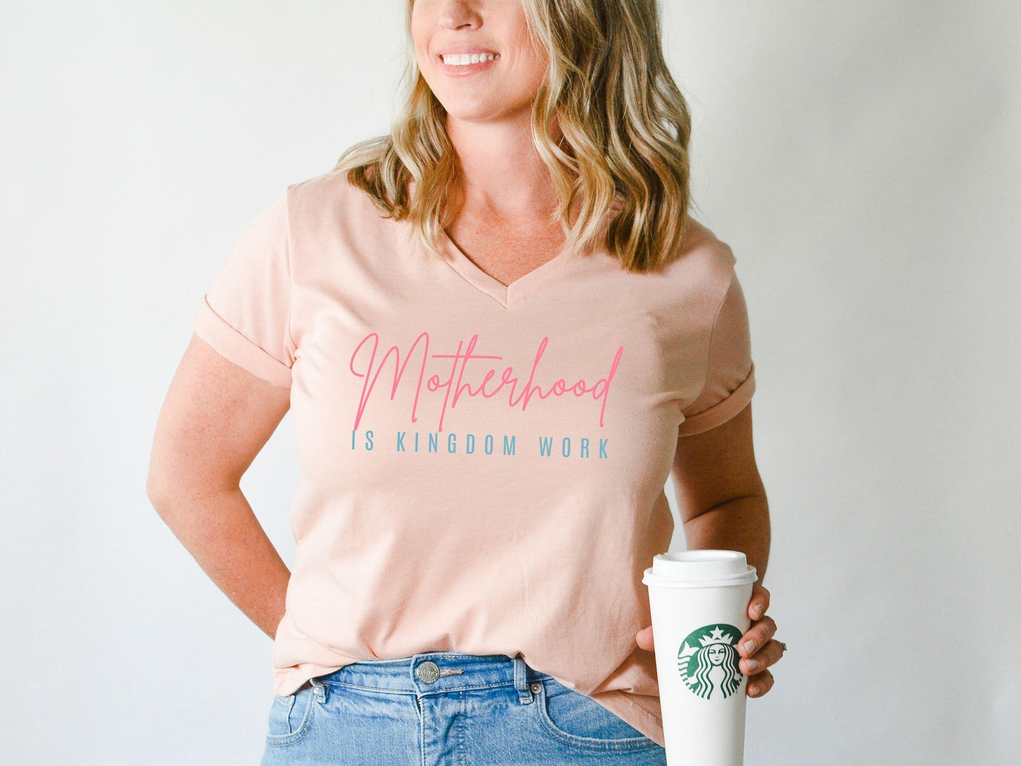 Woman wearing light peach V-neck t-shirt with Motherhood is Kingdom Work design in pink and blue from Abound in Faith