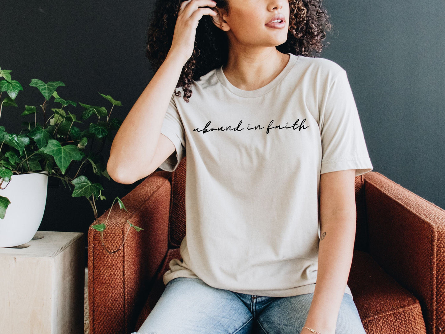 Woman wearing a heather dust Abound in Faith Women’s T-Shirt with elegant script font, seated on a rust-colored chair, looking to the side.