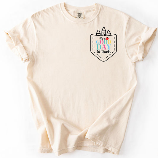 Cream t-shirt with "It’s a Good Day to Teach" printed on left chest pocket with crayon doodles