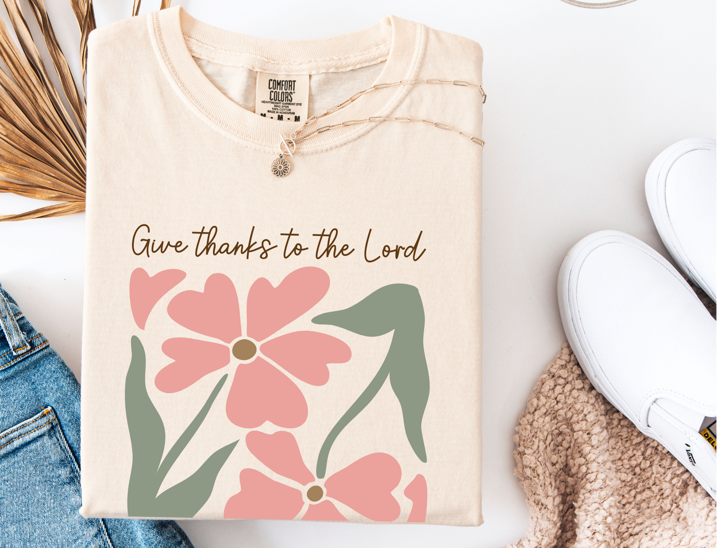 Beige sweatshirt with floral design and 'Give thanks to the Lord' text on a white surface.