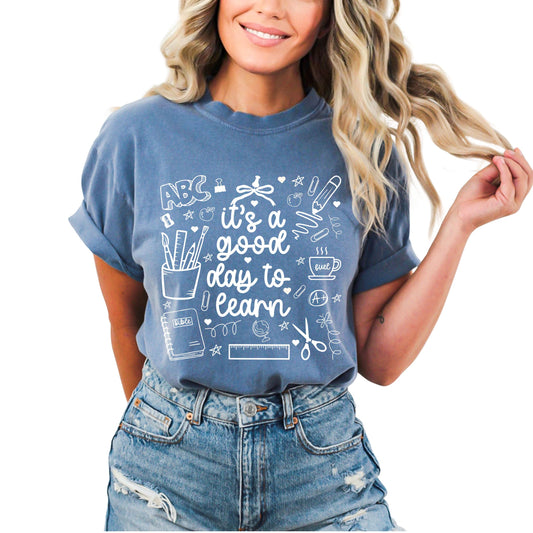 Woman wearing a blue “It’s a Good Day to Learn” graphic teacher t-shirt featuring school doodles and text.